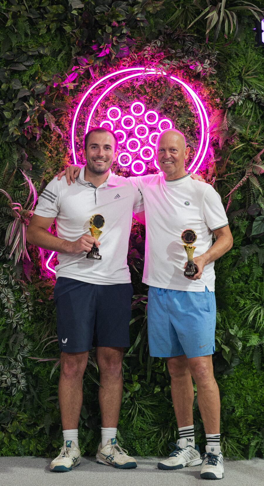 Two players with their arms around each other, each holding a gold trophy, in front of a green wall with a pink neon racket.