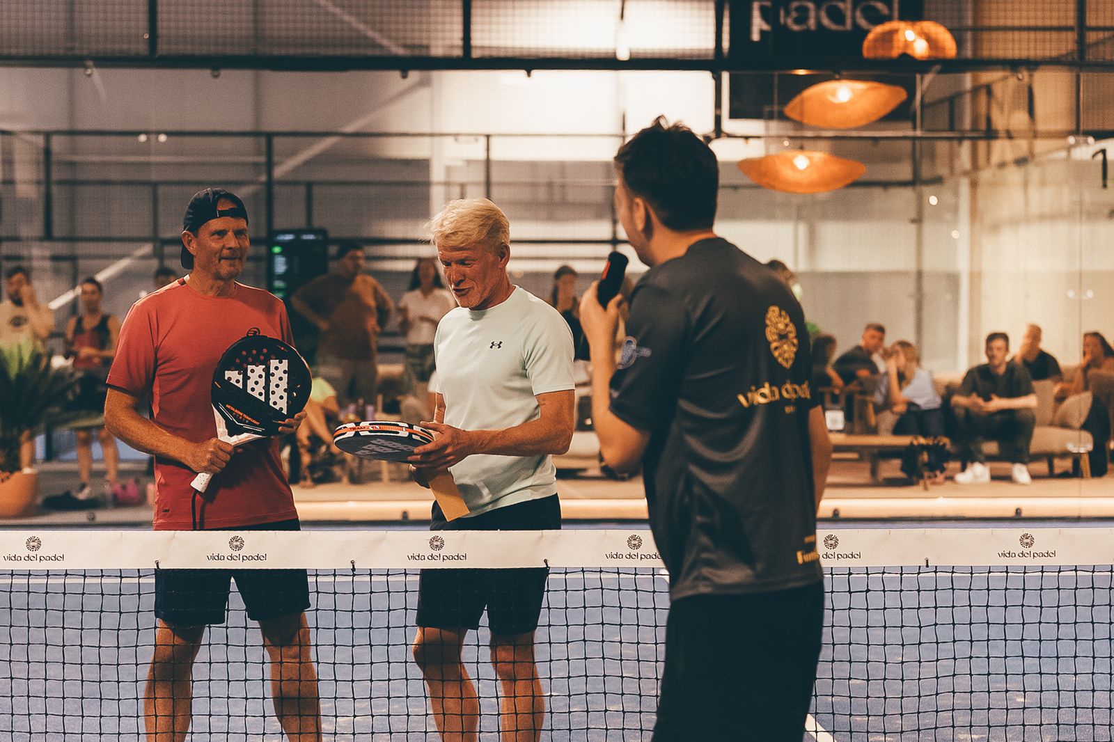 Two spectators at the net, holding rackets, with a host holding a microphone, after winning a challenge at the Vida Del Padel finals.
